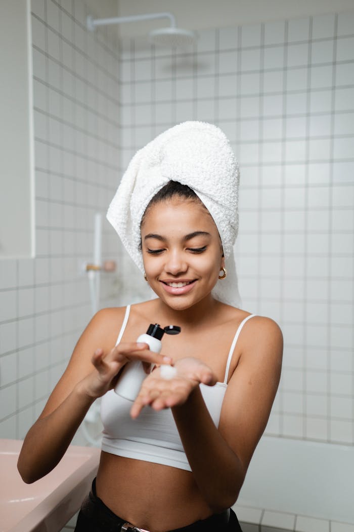 Young woman with towel on head applies lotion as part of her daily skincare routine in a contemporary bathroom.